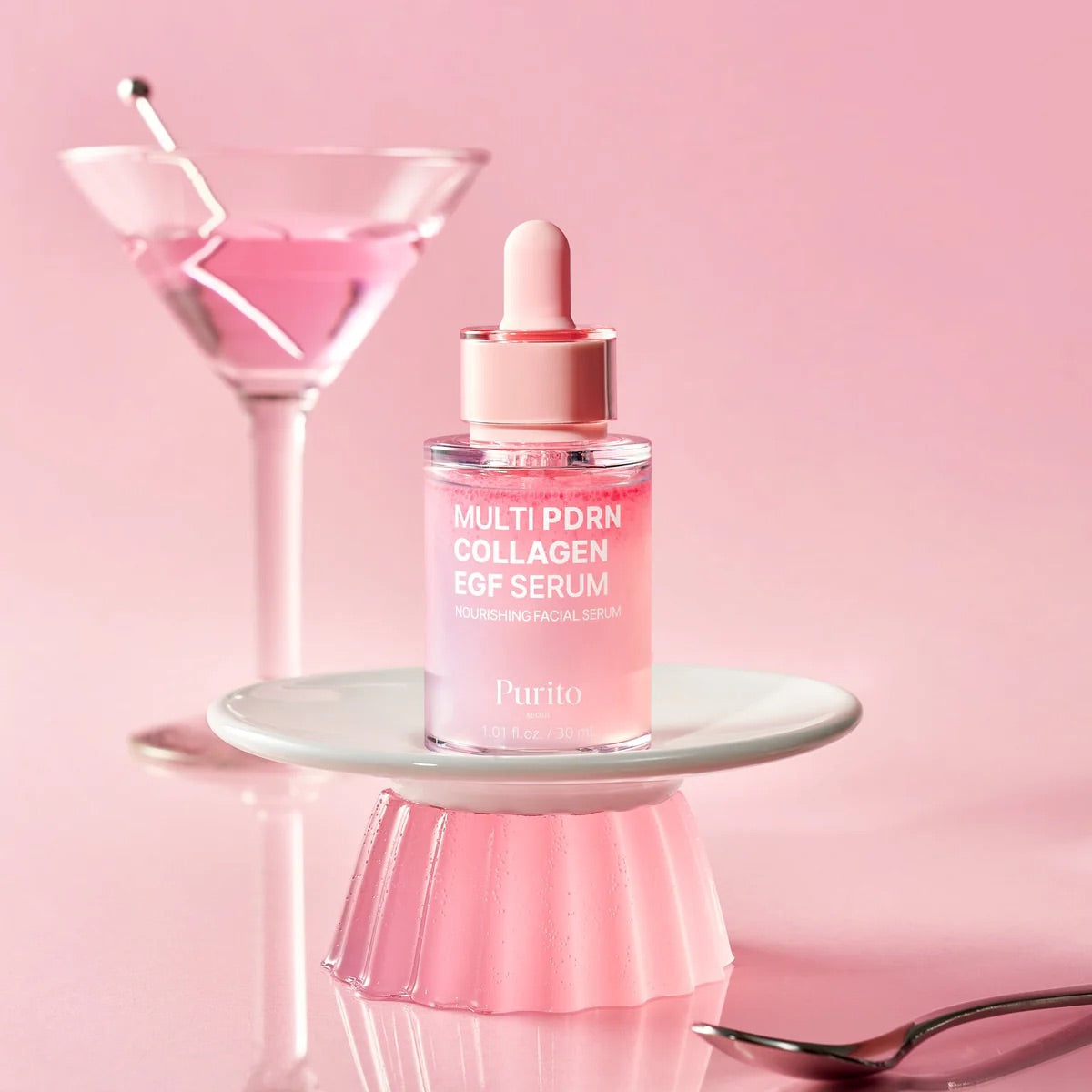 Purito Multi PDRN Collagen EGF Facial Serum in a pink dropper bottle, displayed on a small pink pedestal against a pink background with a pink martini glass and spoon.