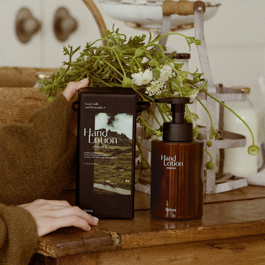 A hand holding a brown box of3. hetras Perfume Essence Hand Lotion 515ml - Flower Shop to a matching amber bottle with a black pump, styled on a rustic wooden table with green foliage and white flowers in the background.