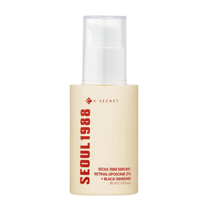 A bottle of K-SECRET SEOUL1988 serum centered on a white background with a soft yellow circular glow behind it. The bottle is cream-colored with black and red text, and the cap is clear and frosted.