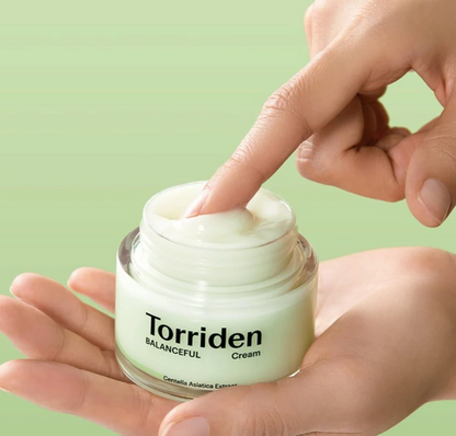 A close-up of a hand holding an open jar of Torriden Balanceful Cream while another hand presses the cream onto the surface. The background is a soft, solid light green.