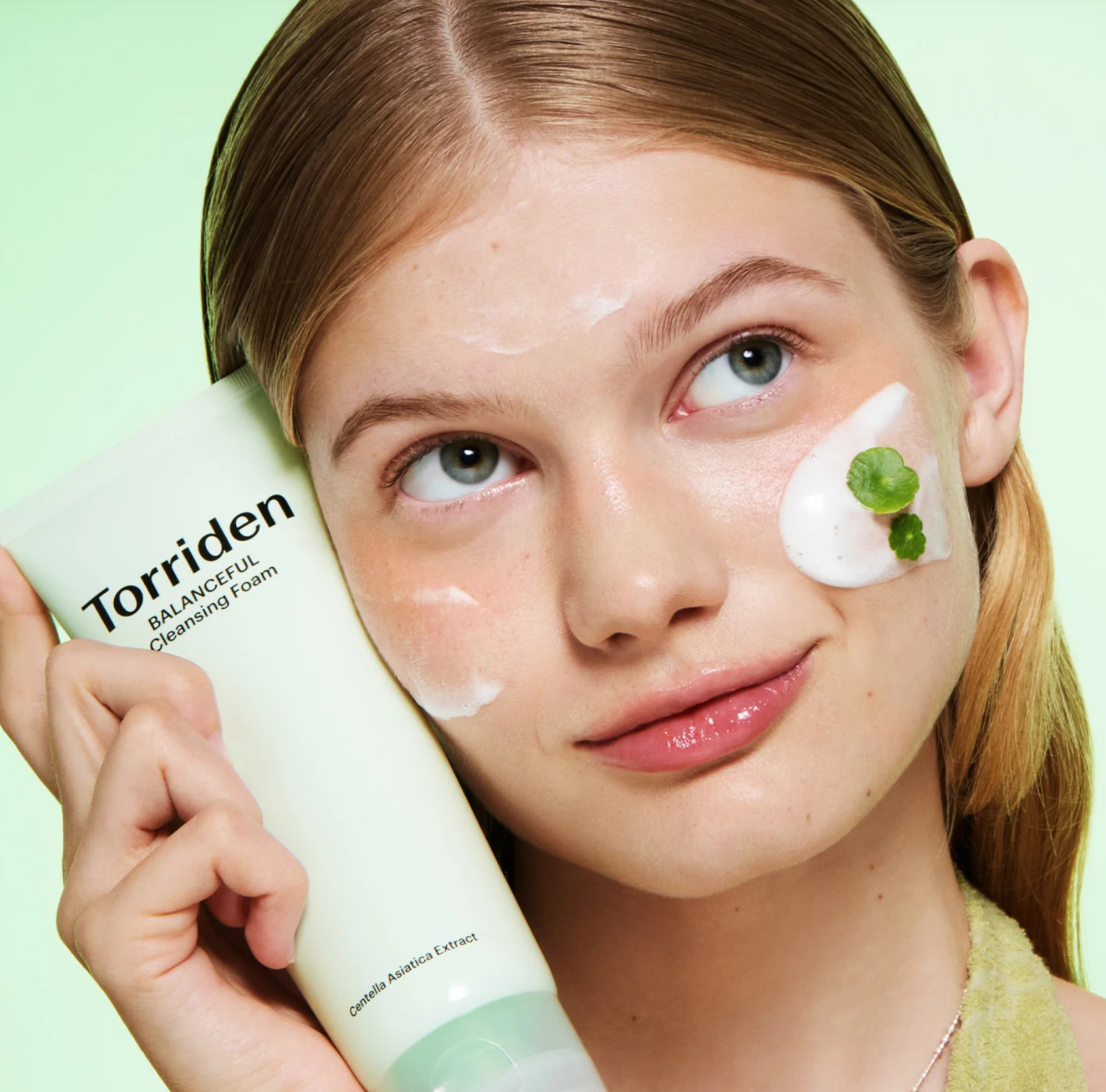 A close-up of a woman applying or demonstrating Torriden Balancing Cleansing F cream, holding the product tube next to her face which has white cream on it, set against a solid pale green background.