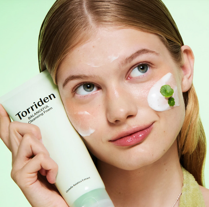 A close-up of a woman applying or demonstrating Torriden Balancing Cleansing F cream, holding the product tube next to her face which has white cream on it, set against a solid pale green background.