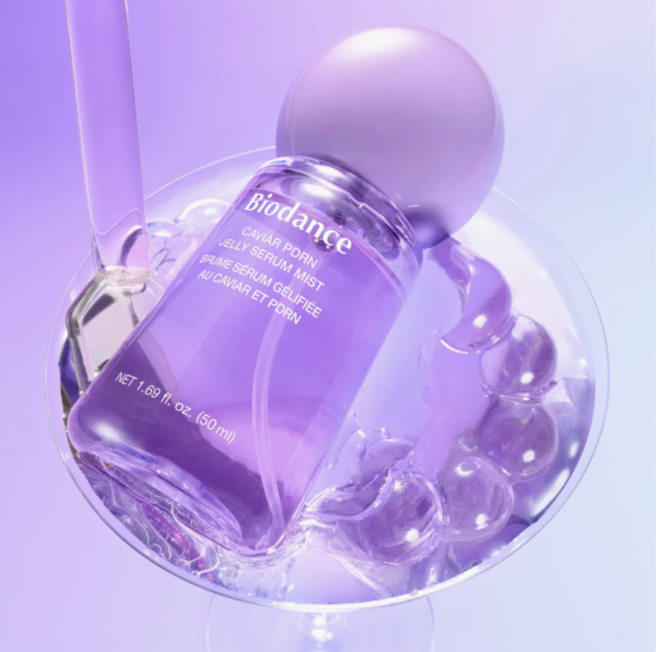 A purple Biodance Caviar Porn Jelly Serum Mist bottle with a matching purple spherical cap, displayed in a glass bowl filled with clear gel and bubble props on a lavender studio background.