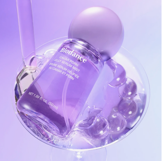 A purple Biodance Caviar Porn Jelly Serum Mist bottle with a matching purple spherical cap, displayed in a glass bowl filled with clear gel and bubble props on a lavender studio background.