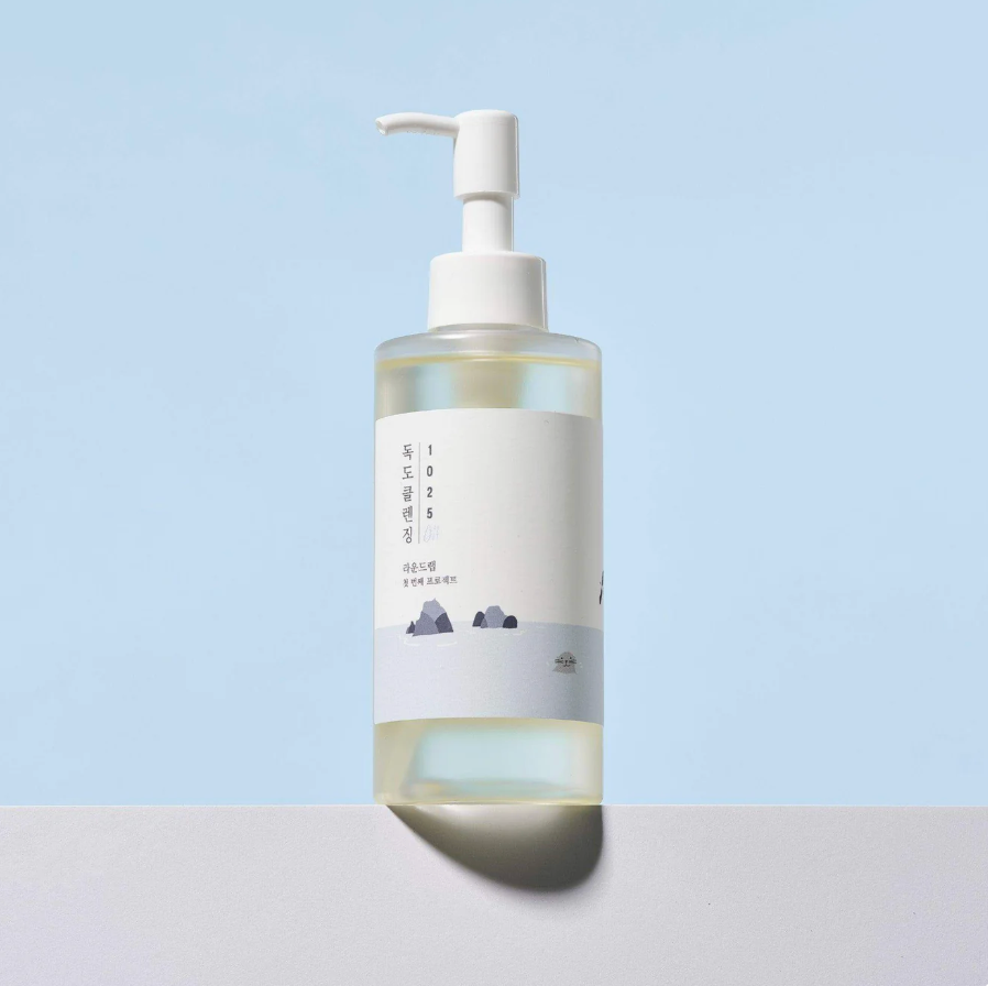 A clear ROUND LAB 1025 Dokdo Cleansing Oil 200ml with a white label and white pump dispenser, containing a pale yellow liquid, standing on a light gray surface against a soft light blue background.