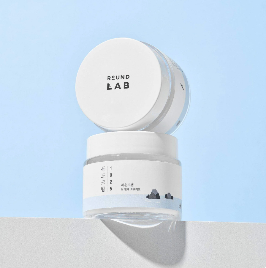 A ROUND LAB 1025 Dokdo Cream 80ml jar with its lid removed and propped against the jar, photographed on a clean white surface against a pale blue background. The product label reads RSUND LAB.